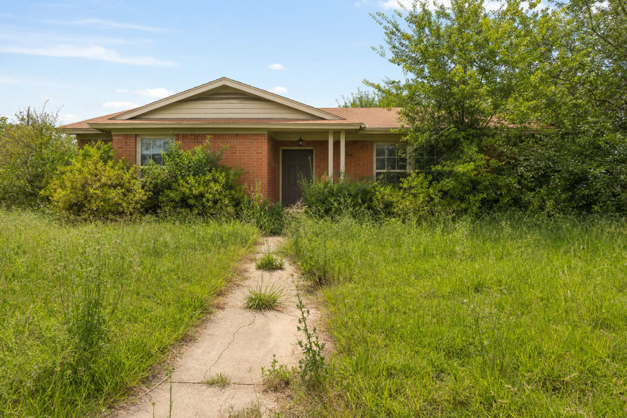 Small brick Texas home with knee-high grass and overgrown shrubs